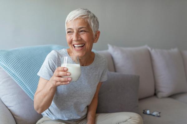 A white woman with short white hair sits on a couch smiling, holding a glass of milk.