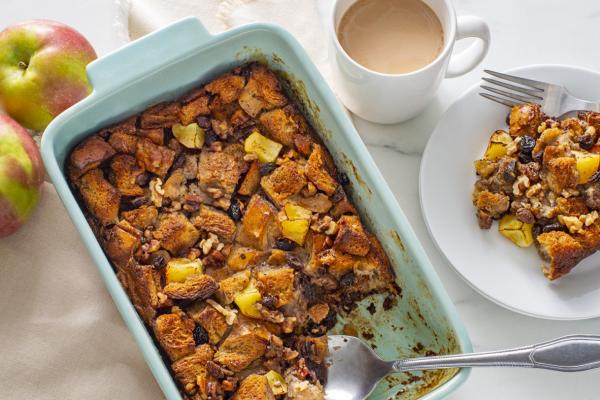 Baked apple raisin bread pudding in a blue ceramic dish, with a serving plated alongside a cup of coffee and fresh apples, ready for a cozy dessert experience.