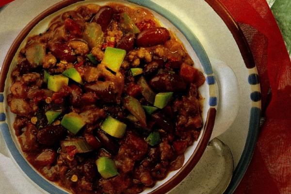 A hearty bowl of Chili Fiesta with chunks of tomato, green bell pepper, and kidney beans, garnished with diced avocado, presented on a rustic plate with blue rim detail.