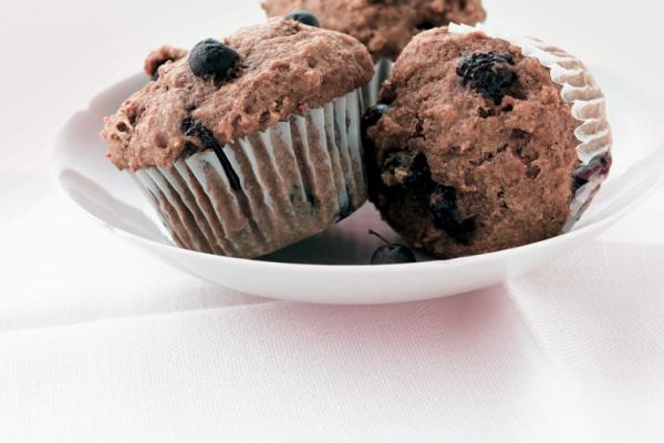 Two blueberry bran muffins in paper liners on a white plate, with fresh blueberries scattered around, on a white textured tablecloth.