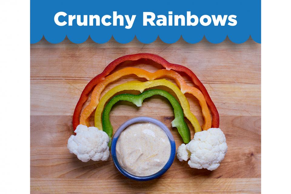 Crunchy Rainbows recipe. Red, orange, yellow and green bell pepper strips arranged in a rainbow shape with two cauliflower florets at each end to make clouds. Yogurt dip in a bowl in the centre. 