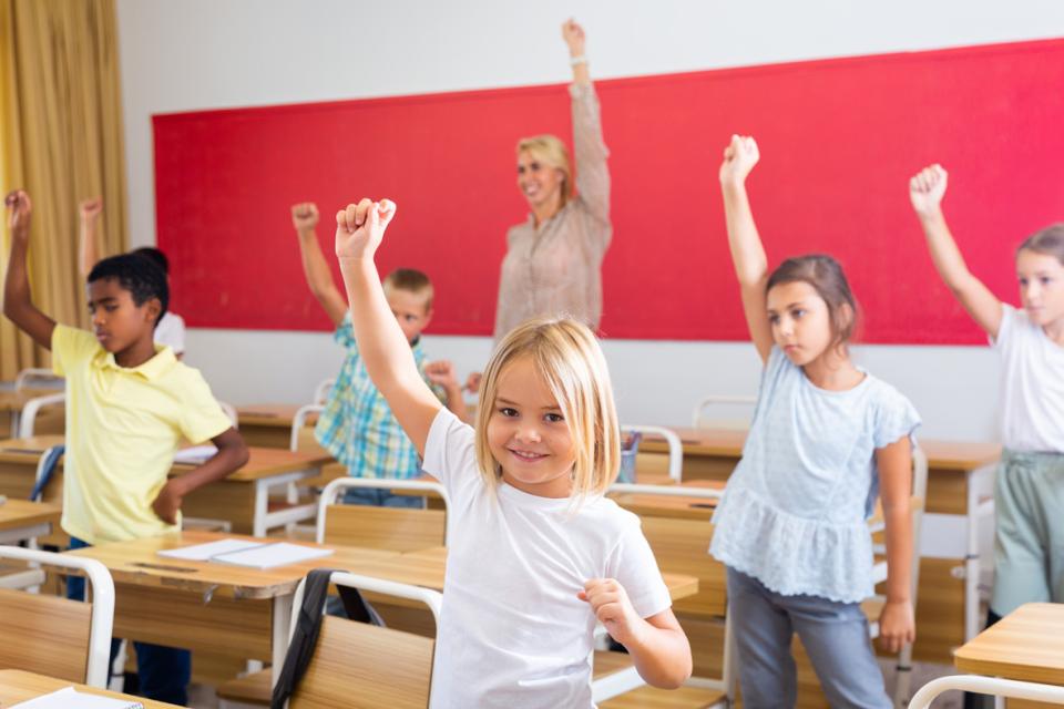 Students standing in a classroom beside their desks doing physical movements