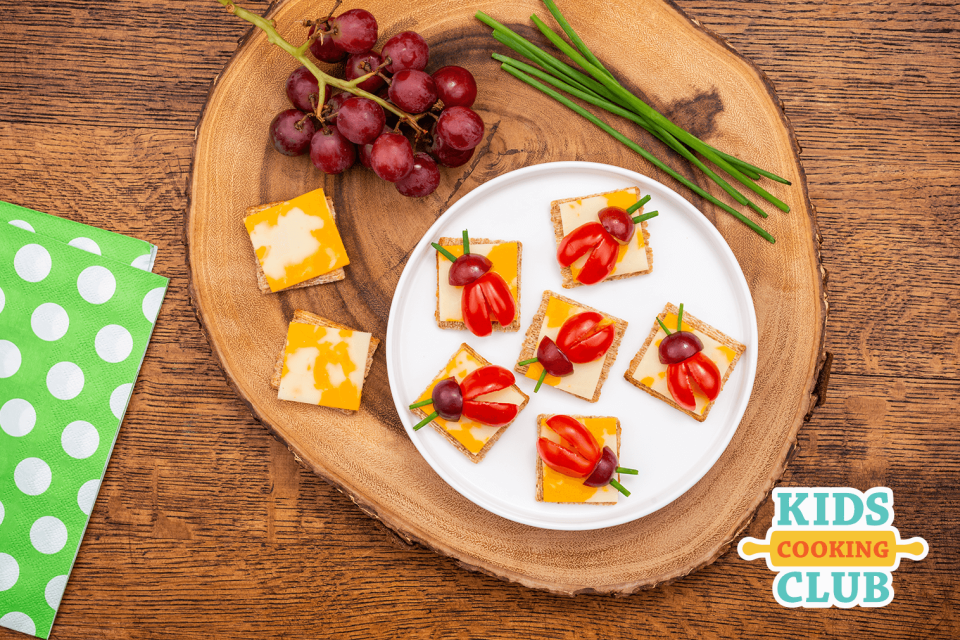 Square crackers and cheese with sliced tomatoes and grapes made to look like a ladybug set on a wooden plank