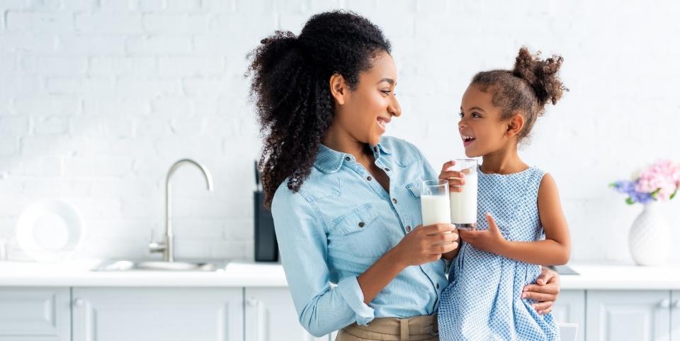 A Black woman with her hair pulled back smiles at a young Black girl beside her as they both hold glasses of milk.