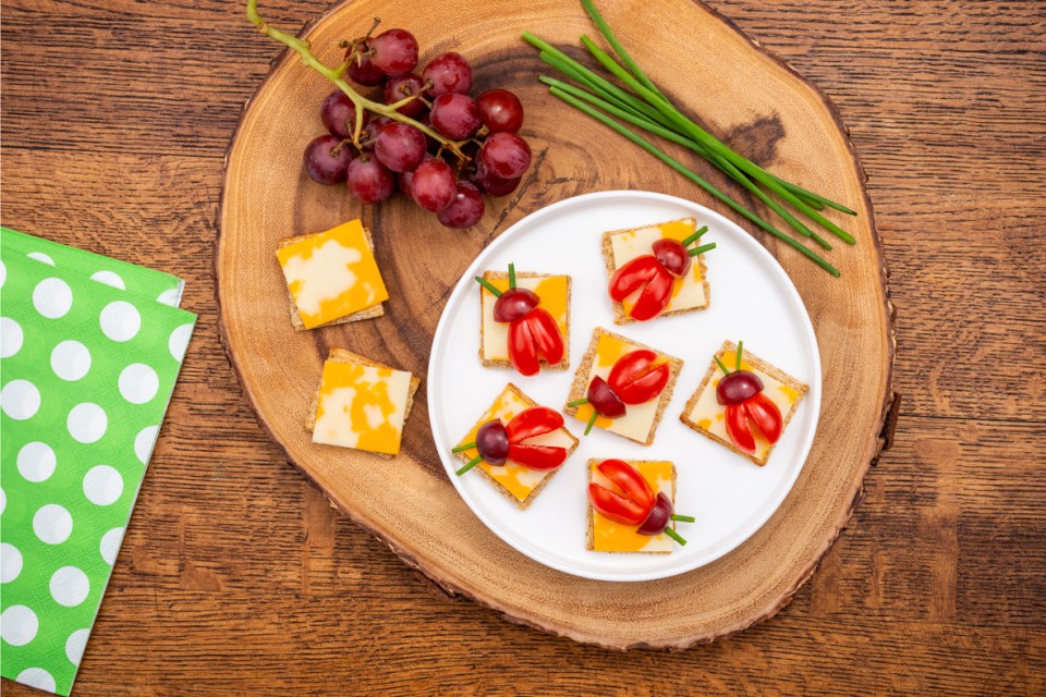 Square cracker and cheese with sliced tomatoes and grapes made to look like a ladybug set on a wooden plank