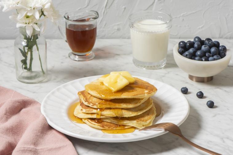 Buttermilk pancakes with syrup and butter, accompanied by milk and berries, set against a backdrop of white flowers on a marble countertop