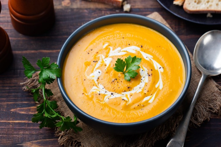 A bowl of creamy carrot squash ginger soup garnished with a swirl of cream and a parsley leaf, served with a spoon on the side, on a rustic wooden table.