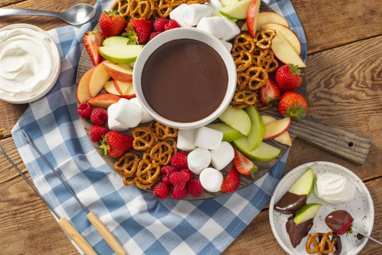 Chocolate fondue in a bowl surrounded by various fruits, marshmallows, and pretzels, ready for dipping