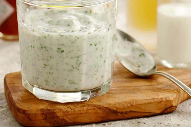 A glass filled with homemade dairy mayonnaise, speckled with finely chopped herbs, resting on a wooden board with a spoon beside it, showcasing a fresh mayonnaise recipe